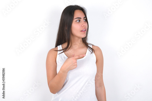 Happy cheerful smiling Young beautiful brunette woman wearing white top over white wall looking and pointing aside with hand. Copy space and advertisement concept. © Jihan
