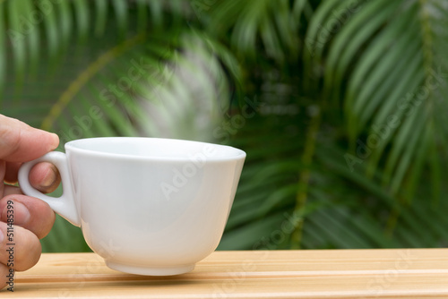 Man hand holding a white coffee mug with stream on a wooden table and palm tree in background.