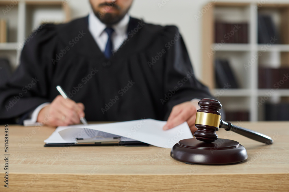 Close up of a gavel on a wooden table in the courtroom, with a male judge putting his signature on some legal documents in the background. Court trial, justice, legislation, law enforcement concepts Close up of a gavel on a wooden table in the courtroom, with a male judge putting his signature on some legal documents in the background. Court trial, justice, legislation, law enforcement concepts