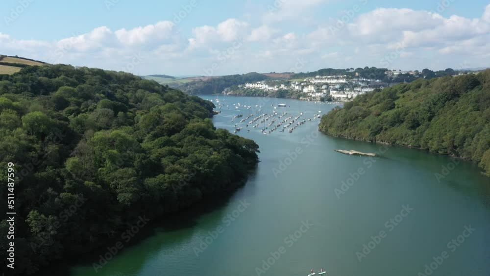 Dramatic wide rising aerial reveal from paddle boarders on the river Fowey, to reveal the Cornish towns of Fowey and Polruan on the coast of Cornwall, UK Dramatic wide rising aerial reveal from paddle boarders on the river Fowey, to reveal the Cornish towns of Fowey and Polruan on the coast of Cornwall, UK
