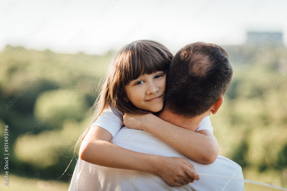 Young girl hugs her father, putting arms around his neck and looking at the camera. Outdoor family walk Young girl hugs her father, putting arms around his neck and looking at the camera. Outdoor family walk