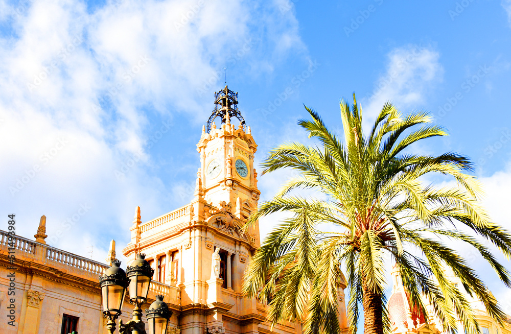 Clock tower building. Large clock tower with hour hands that shows time. Palm trees on the background of the facade of a residential building. Architecture of modern buildings in Valencia, Spain.