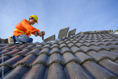 A male roofing installer is working on the roof of a house. Construction workers repair the roof of a house using drills and screws.