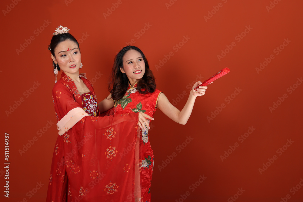 two young attractive Asian woman wearing tadeonal Chinese red hanfu long skirt scarf and cheongsam costume dress hand fan on read maroon background wall pose look talk point show
