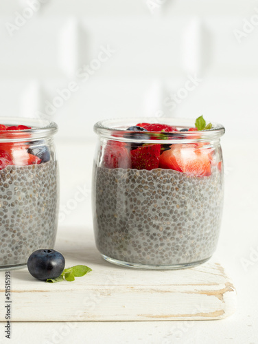 Vegan chia pudding with coconut milk, strawberry, blueberry and jerusalem artichoke syrup in the glass jars on the white wooden serving desk. Easy summer dessert. White background, high key, close up