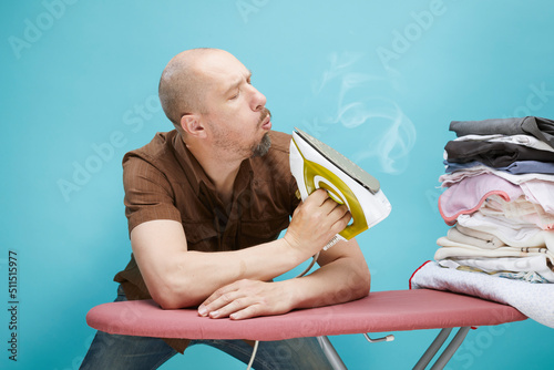 A funny bald man with emotions using electrical iron on ironing board over on blue background.