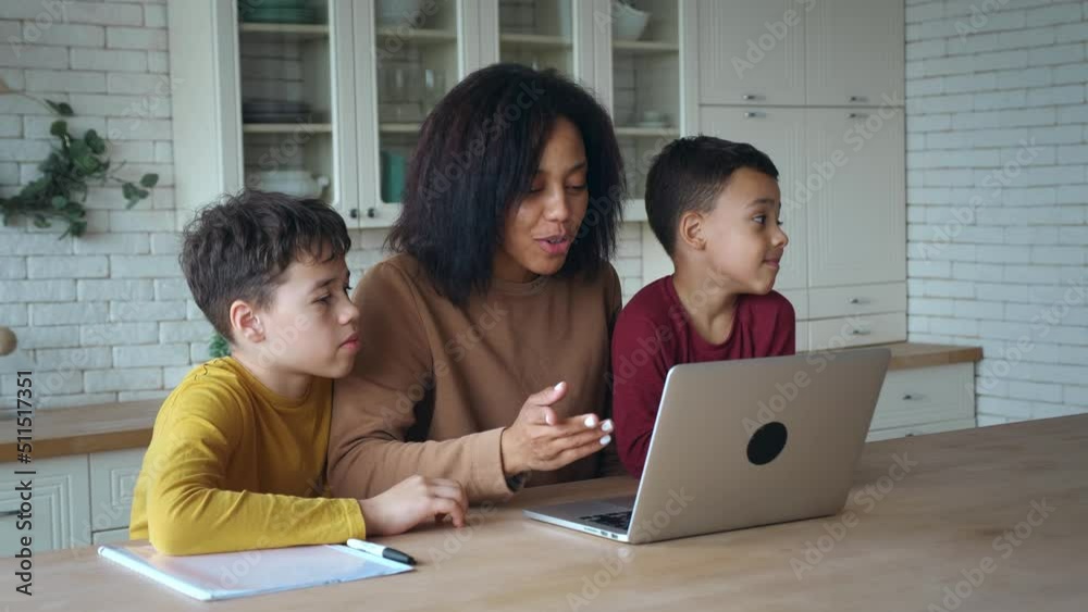 African American woman and sons sitting at the table in the kitchen ,waving hands looking at web camera using laptop for video call. Smiling mom and children boys greeting online making video call African American woman and sons sitting at the table in the kitchen ,waving hands looking at web camera using laptop for video call. Smiling mom and children boys greeting online making video call