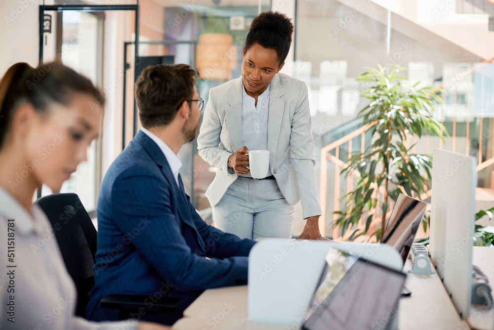 Happy black businesswoman talks to her colleague who is working on laptop in office. Happy black businesswoman talks to her colleague who is working on laptop in office.