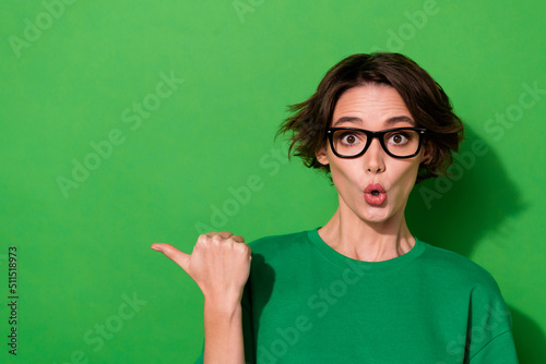 Photo of excited funky young lady dressed casual t-shirt eyewear pointing thumb empty space isolated green color background
