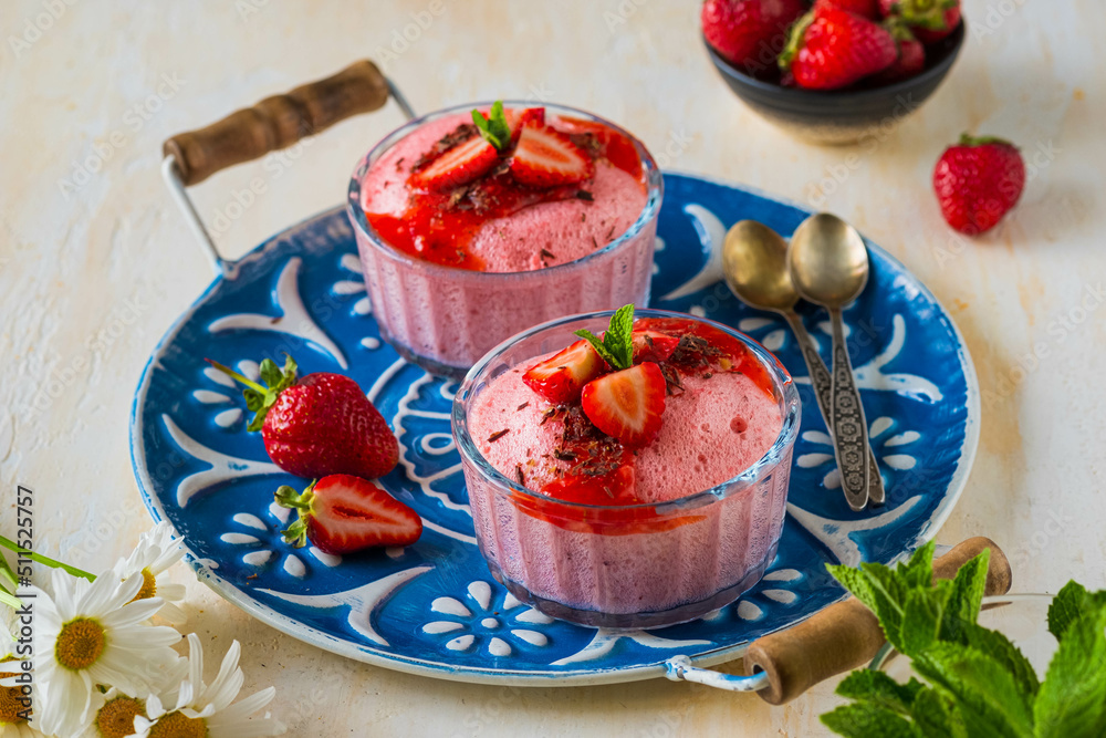 Dessert, airy strawberry mousse in glass bowls, decorated with fresh strawberries, syrup and chocolate chips on a blue tray on a light concrete background.