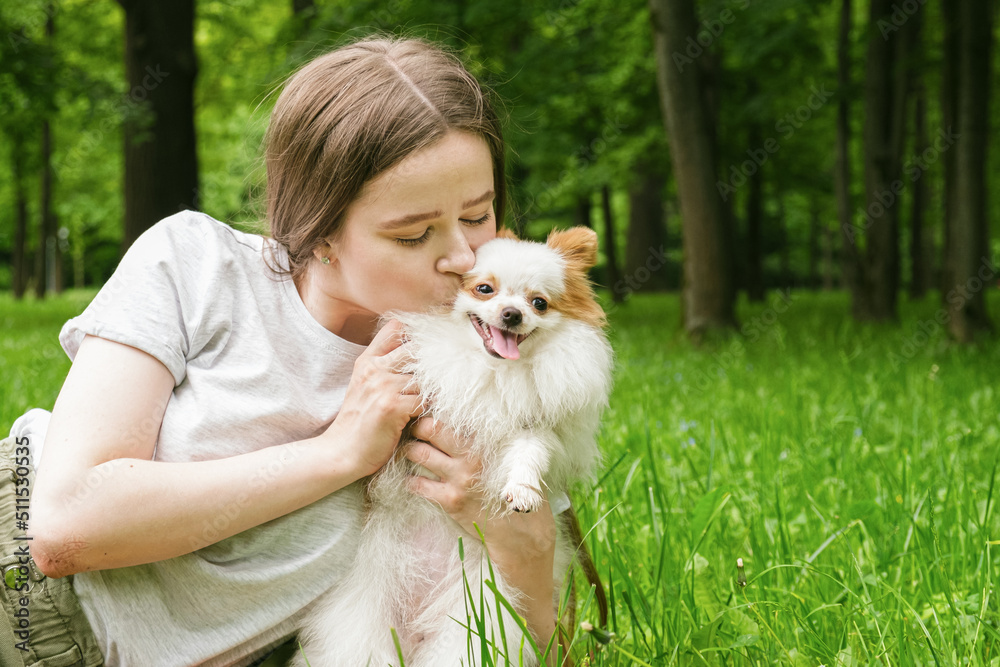 Young woman with long hair lies on the lawn in the park with a Pomeranian dog. Funny fluffy dog with white fur and red ears stuck out his tongue. Woman kisses a dog. Space for text. Young woman with long hair lies on the lawn in the park with a Pomeranian dog. Funny fluffy dog with white fur and red ears stuck out his tongue. Woman kisses a dog. Space for text.