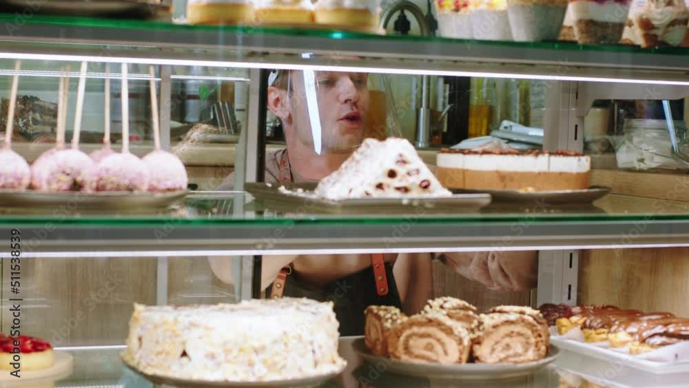 In the modern bakery cafe salesman with protective mask very charismatic arrange the all deserts from the showcase fridge very carefully In the modern bakery cafe salesman with protective mask very charismatic arrange the all deserts from the showcase fridge very carefully