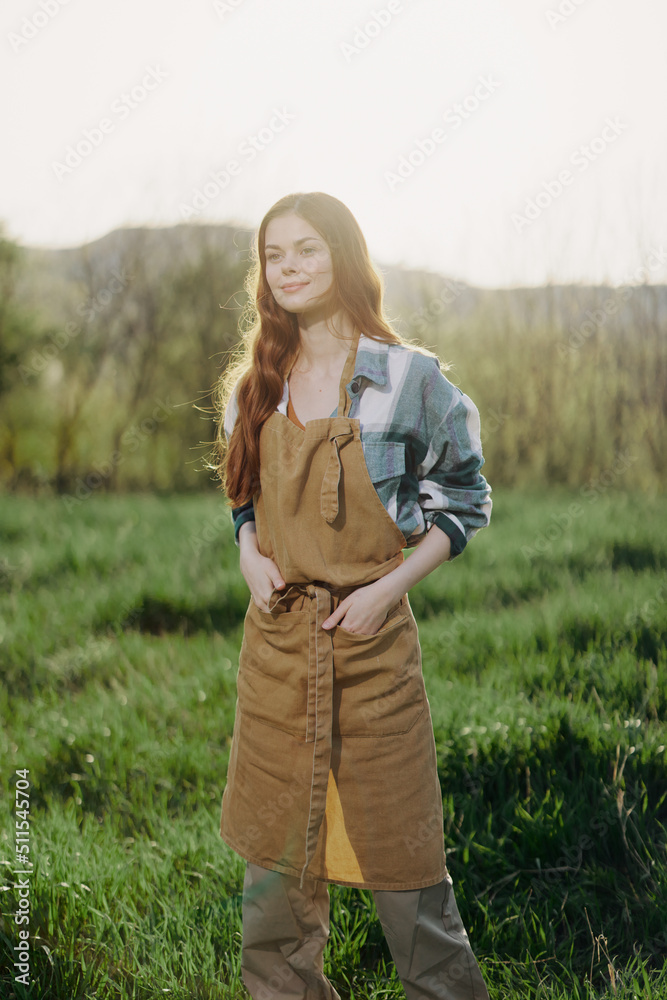 A farmer woman in her work clothes, plaid shirt and apron, stands in the field on the green grass and smiles in the setting sun after a hard day's work A farmer woman in her work clothes, plaid shirt and apron, stands in the field on the green grass and smiles in the setting sun after a hard day's work