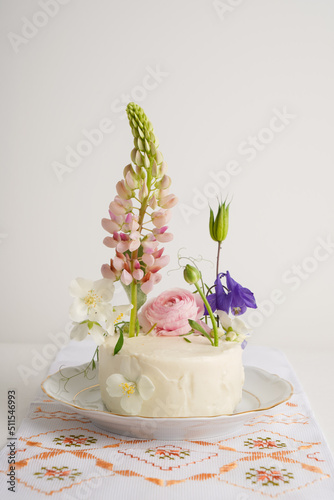 Tiny wedding cake with fresh flowers with lupine, on a light background with local confectionery. Vegan wedding white cake on light background