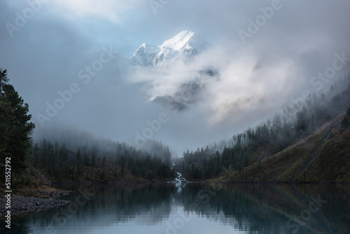 Fototapeta Naklejka Na Ścianę i Meble -  Tranquil scenery with snow castle in clouds. Mountain creek flows from forest hills into glacial lake. Snowy mountains in fog clearance. Small river and coniferous trees reflected in calm alpine lake.