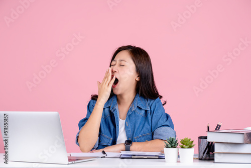 Tired Asian woman wearing casual shirt sit work at white office desk. she is yawning isolated on a pink background