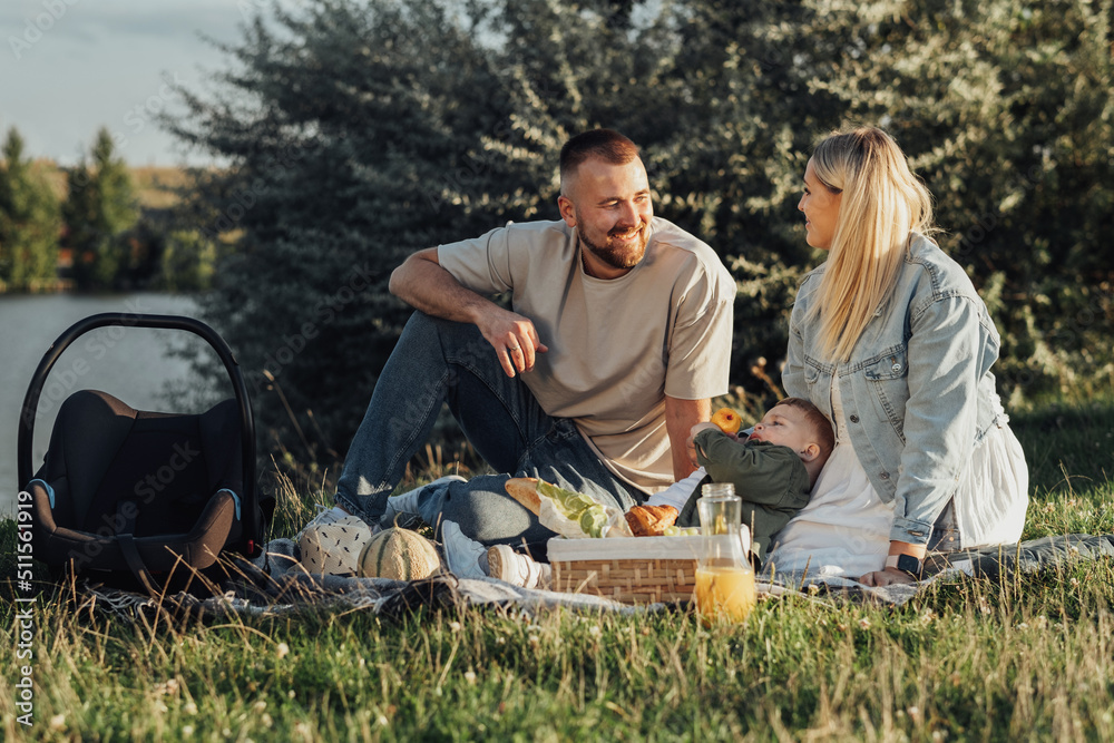 Happy Couple with Their Toddler Son Having a Picnic Outdoors by the Lake, Young Family Weekend Concept Happy Couple with Their Toddler Son Having a Picnic Outdoors by the Lake, Young Family Weekend Concept