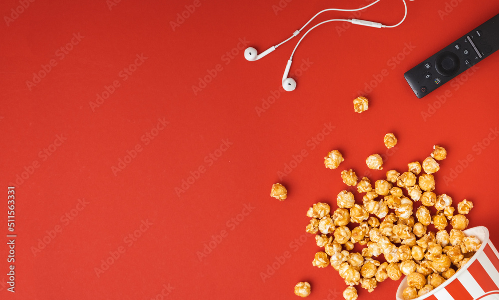 Fototapeta premium Caramel popcorn scattered from a red and white bucket with remote control and headphones on a red background. Flat lay. cinema advertising