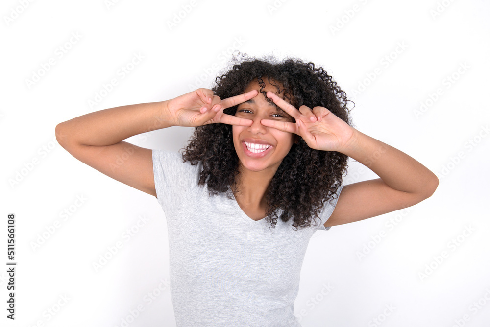 Cheerful positive Young beautiful girl with afro hairstyle wearing grey t-shirt over white wall shows v-sign near eyes open mouth Cheerful positive Young beautiful girl with afro hairstyle wearing grey t-shirt over white wall shows v-sign near eyes open mouth