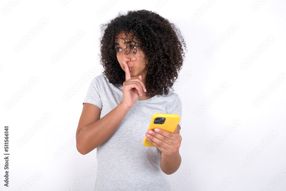 Young beautiful girl with afro hairstyle wearing gray t-shirt over white background holding modern gadget ask not tell secrets Young beautiful girl with afro hairstyle wearing gray t-shirt over white background holding modern gadget ask not tell secrets