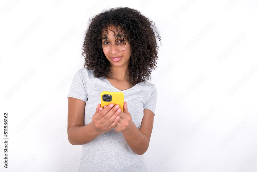 Portrait of serious confident Young beautiful girl with afro hairstyle wearing gray t-shirt over white background holding phone in two hands