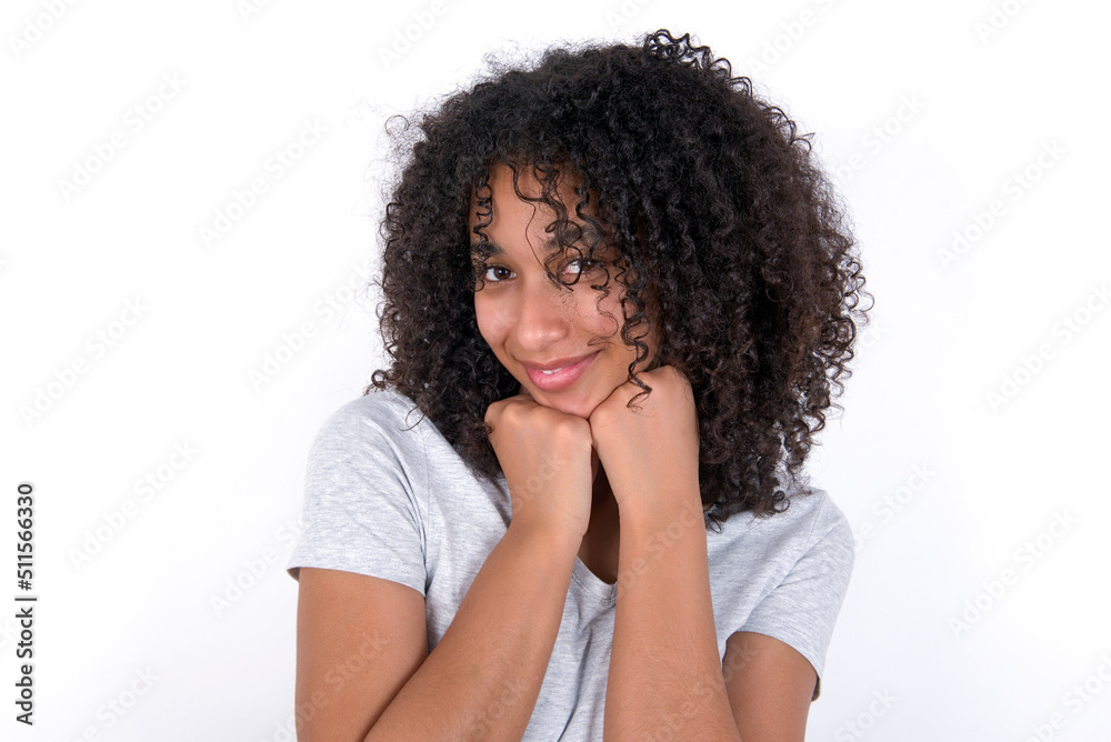 Satisfied Young beautiful girl with afro hairstyle wearing gray t-shirt over white background touches chin with both hands, smiles pleasantly, rejoices good day with lover Satisfied Young beautiful girl with afro hairstyle wearing gray t-shirt over white background touches chin with both hands, smiles pleasantly, rejoices good day with lover