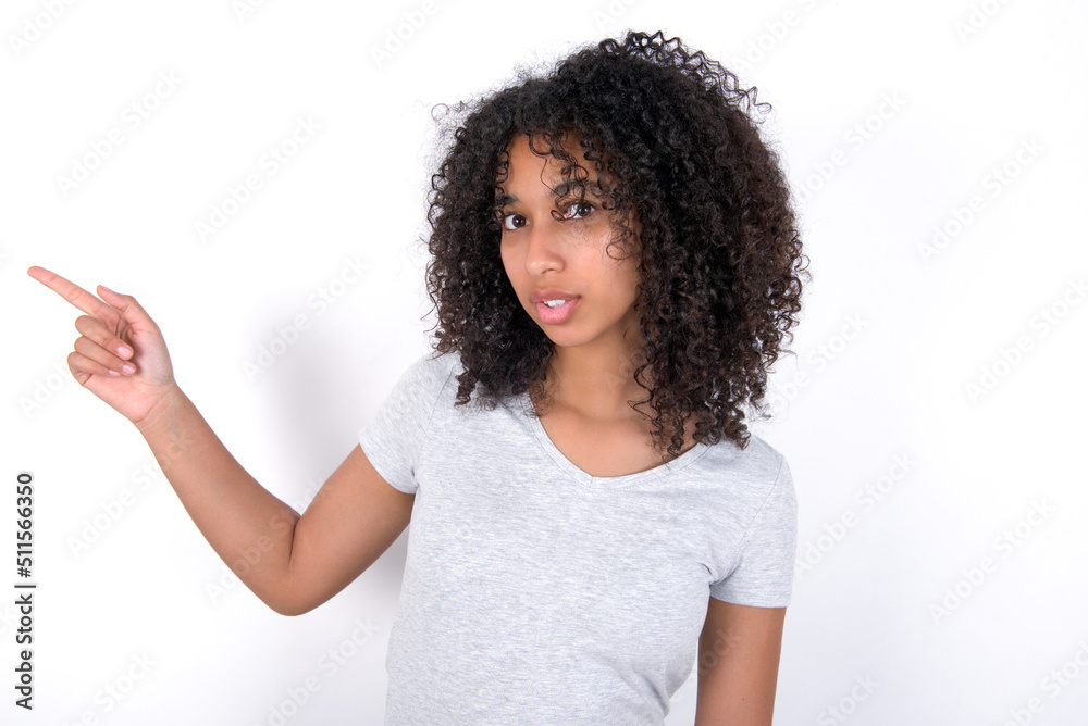 Emotive Young beautiful girl with afro hairstyle wearing gray t-shirt keeps jaw dropped from shock demonstrates amazing promo points right on blank space demonstrates big shopping sale. Advertisement