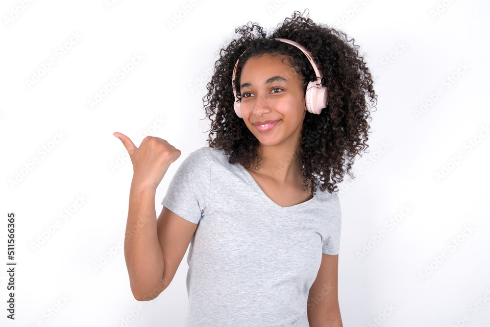 Young beautiful girl with afro hairstyle wearing gray t-shirt over white background listens audio track via wireless headphones points thumb away advertises copy space Young beautiful girl with afro hairstyle wearing gray t-shirt over white background listens audio track via wireless headphones points thumb away advertises copy space