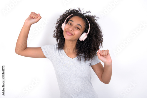 Carefree Young beautiful girl with afro hairstyle wearing gray t-shirt over white with toothy smile raises arms dances carefree moves with rhythm of music listens music from playlist via headphones