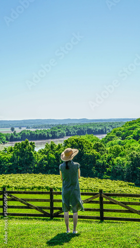 Woman in sunhat standing in a vineyard overlooking a valley © Reed