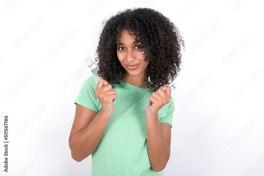 hungry Young beautiful girl with afro hairstyle wearing green t-shirt over white wall holding in hand fork knife want tasty yummy pizza pie hungry Young beautiful girl with afro hairstyle wearing green t-shirt over white wall holding in hand fork knife want tasty yummy pizza pie