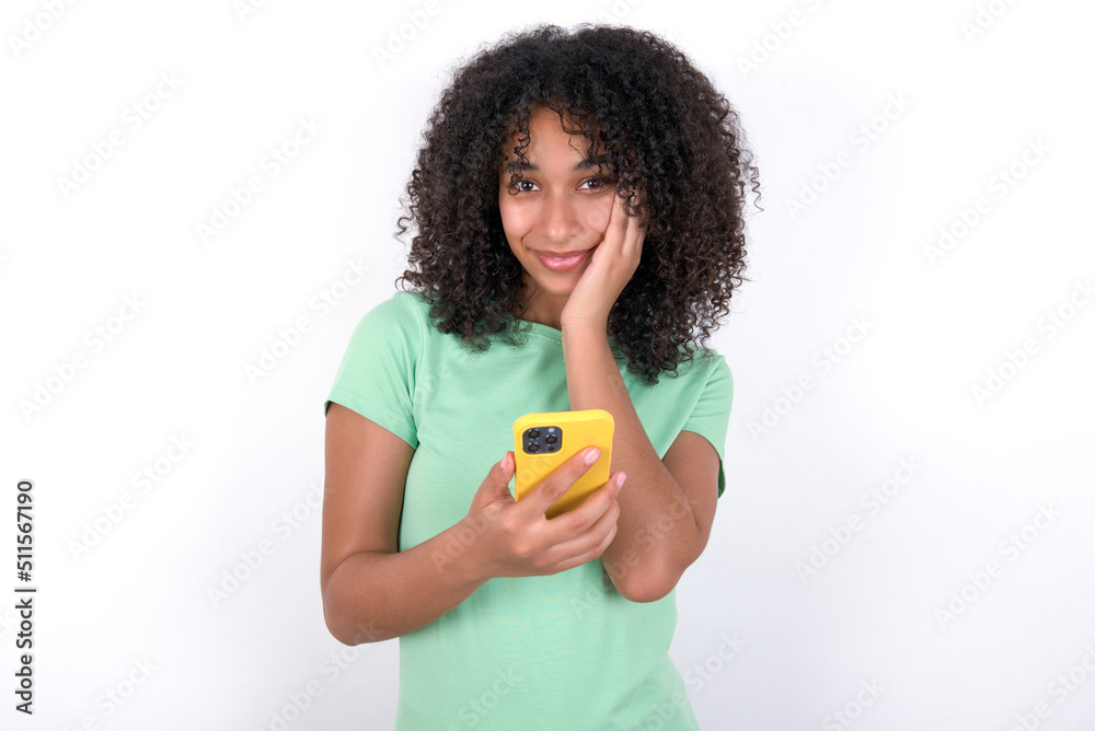 Joyous Young beautiful girl with afro hairstyle wearing green t-shirt over white wall poses with mobile phone device, types text message on modern smartphone, watches funny video during free time, Joyous Young beautiful girl with afro hairstyle wearing green t-shirt over white wall poses with mobile phone device, types text message on modern smartphone, watches funny video during free time,