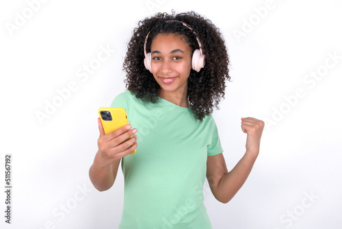 Positive Young beautiful girl with afro hairstyle wearing green t-shirt over white background holds modern cell phone connected to headphones, clenches fist from good emotions, exclaims with joy,