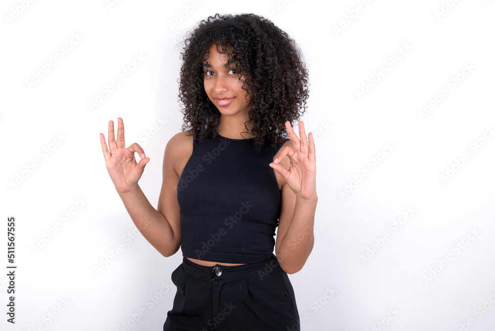 Young beautiful girl with afro hairstyle wearing black tank top over white background showing both hands with fingers in OK sign. Approval or recommending concept Young beautiful girl with afro hairstyle wearing black tank top over white background showing both hands with fingers in OK sign. Approval or recommending concept