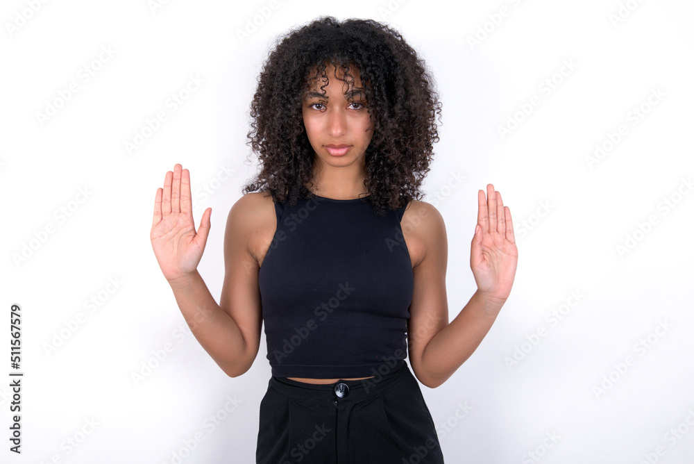 Serious Young beautiful girl with afro hairstyle wearing black tank top over white background pulls palms towards camera, makes stop gesture, asks to control your emotions and not be nervous Serious Young beautiful girl with afro hairstyle wearing black tank top over white background pulls palms towards camera, makes stop gesture, asks to control your emotions and not be nervous