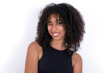 Coquettish Young beautiful girl with afro hairstyle wearing black tank top over white background smiling happily, blinking at camera in a playful manner, flirting with you.