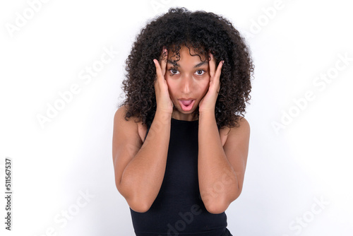 Young beautiful girl with afro hairstyle wearing black tank top over white background with scared expression, keeps hands on head, jaw dropped, has terrific expression. Omg concept