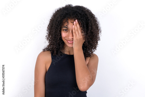 Young beautiful girl with afro hairstyle wearing black tank top over white background covering one eye with her hand, confident smile on face and surprise emotion.