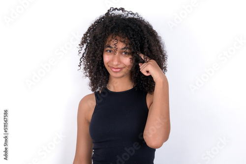 Young beautiful girl with afro hairstyle wearing black tank top over white background tries to memorize something, keeps fore finger on temple, reminds information for exam, © Roquillo