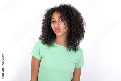 Shot of pleasant looking Young beautiful girl with afro hairstyle wearing green t-shirt over white background , pouts lips, looks at camera, Human facial expressions