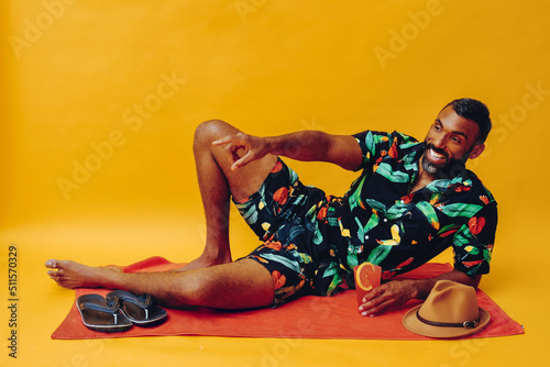 handsome bearded mid adult african american man smiling on vacation lying on an orange towel, holding orange juice looking away studio shot