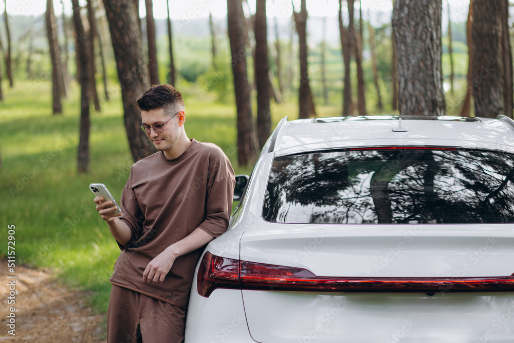 A young guy with a beard talking on the phone near his electric car. In his hands he has a fashionable smartphone. He has a good mood.