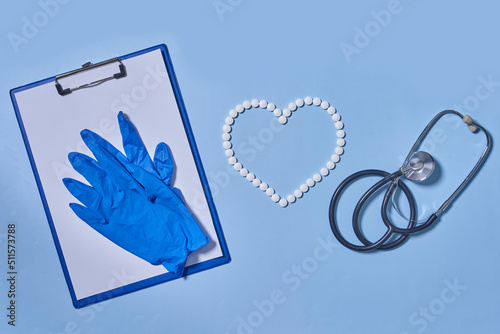 Group of white round pills forms heart figure from white pills, notebook, blue gloves and stethoscopen on blue background. Theme of health care, medical treatment and disease prevention. photo