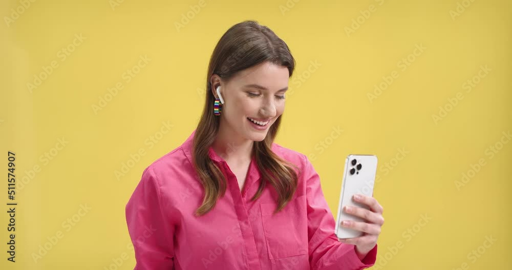 Joyful beautiful young female in pink blouse standing on yellow background in studio waving hand and talking on video chat on smartphone in wireless earphones. Video call, communication, close up Joyful beautiful young female in pink blouse standing on yellow background in studio waving hand and talking on video chat on smartphone in wireless earphones. Video call, communication, close up
