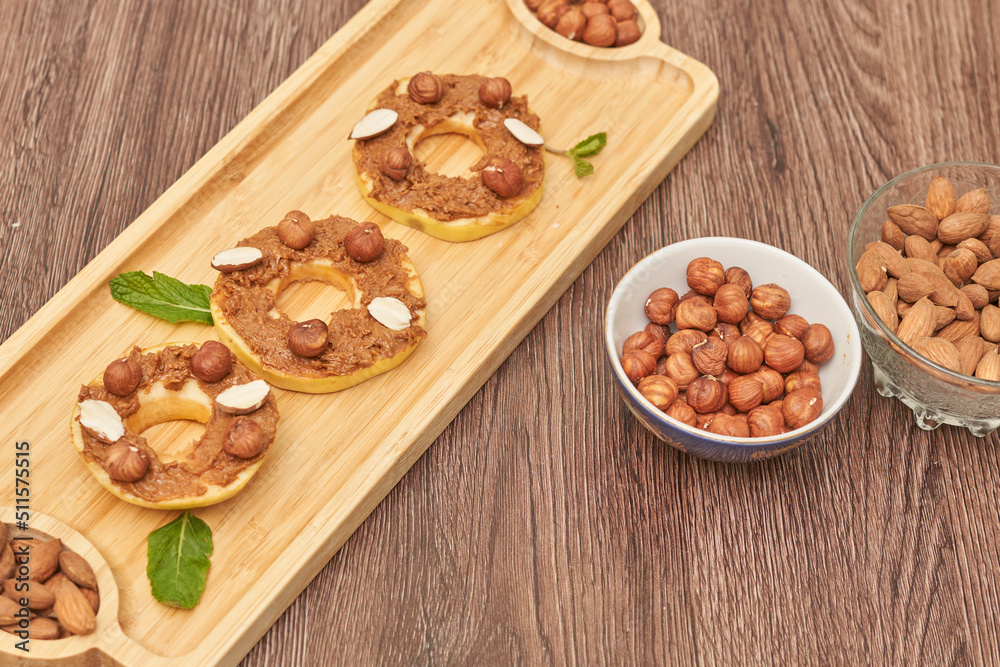 Fototapeta premium Apple rounds dialed on top of each other with peanut butter, almonds and nuts, on wood plate on the table. Healthy food