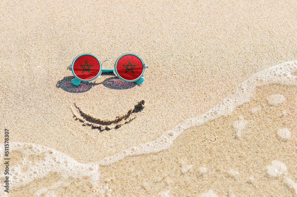 Obraz premium A painted smile on the beach and sunglasses with the flag of Morocco. The concept of a positive holiday in the resort of Morocco.
