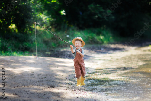 Preschooler boy in yellow rubber boots plays fishing, child fishes with stick near lake in countryside, games in nature and childhood without gadgets, summer holidays