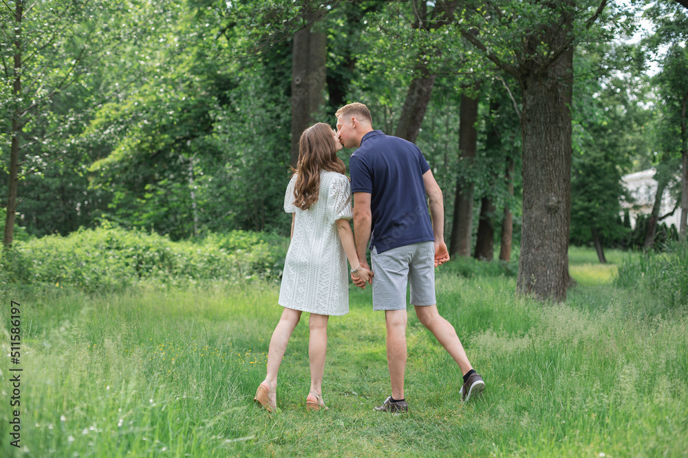 Side back view full length portrait of passionate romantic young couple kissing, holding hands in summer park