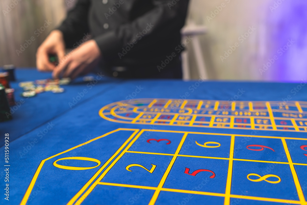 Naklejka premium Vibrant casino table with roulette in motion, with casino chips, tokens, the hand of croupier, dollar bill money and a group of gambling rich wealthy people playing bet in the background