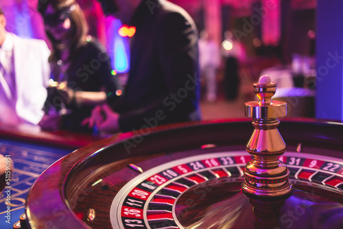 Vibrant casino table with roulette in motion, with casino chips, tokens, the hand of croupier, dollar bill money and a group of gambling rich wealthy people playing bet in the background